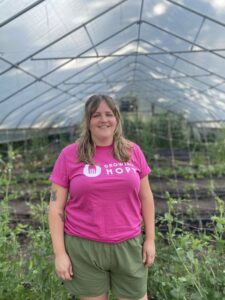 Jennifer Pritchett wearing a pink Growing Hope Shirt in a hoop house with many plants behind her. 
